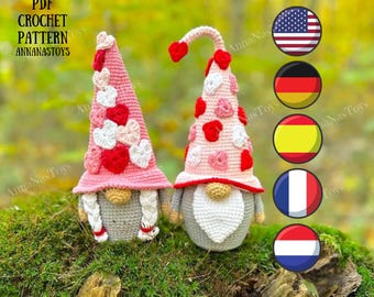 May include: Two crocheted gnomes with pink and white heart hats. The gnomes are sitting on a bed of green moss. The image includes the text "PDF CROCHET PATTERN ANNANASTOYS".