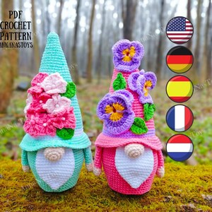 May include: Two crocheted gnomes with floral accents. One gnome is wearing a teal hat with pink flowers and the other is wearing a pink hat with purple flowers. The gnomes are sitting on a bed of moss.