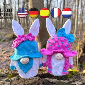 May include: Two crocheted Easter bunny gnomes, one wearing a blue hat with pink flowers and the other wearing a pink hat with blue polka dots. Both gnomes have white yarn hair and white yarn bodies.