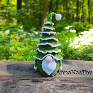 Puede incluir: Un gnomo de &aacute;rbol de Navidad de crochet verde con barba blanca y gorro blanco con pomp&oacute;n. El gnomo est&aacute; decorado con cuentas de colores y est&aacute; sentado sobre una superficie de madera. AnnaNasToys est&aacute; escrito en la superficie.