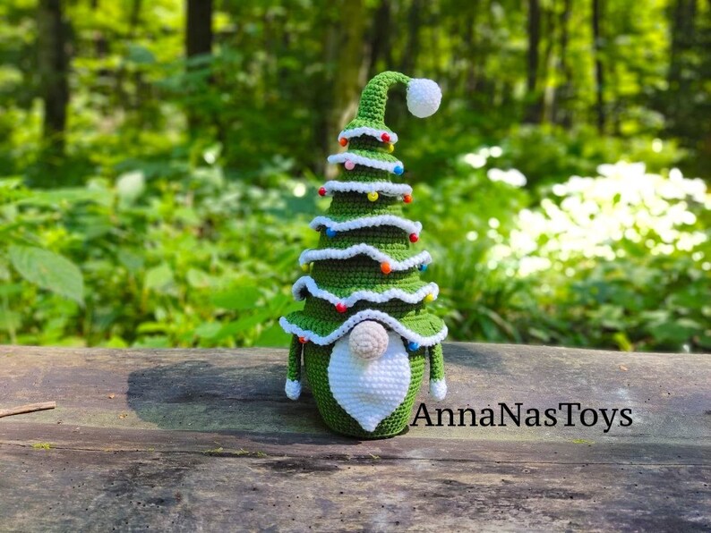 Puede incluir: Un gnomo de &aacute;rbol de Navidad de crochet verde con barba blanca y gorro blanco con pomp&oacute;n. El gnomo est&aacute; decorado con cuentas de colores y est&aacute; sentado sobre una superficie de madera. AnnaNasToys est&aacute; escrito en la superficie.