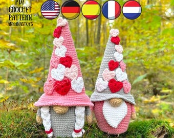 May include: Two crocheted gnomes with pink and white hearts on their hats. The gnomes are standing on a bed of moss. The gnomes are wearing grey pants and white braids. The image includes the text "PDF CROCHET PATTERN ANNANASTOYS".