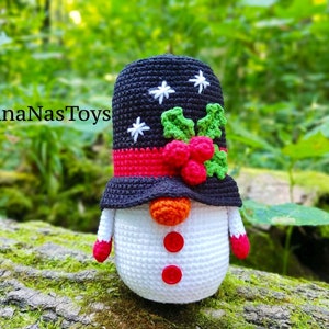 May include: A crocheted snowman ornament with a black hat, red and green holly, and white snowflakes. The snowman is wearing a red scarf and has an orange carrot nose and red buttons.