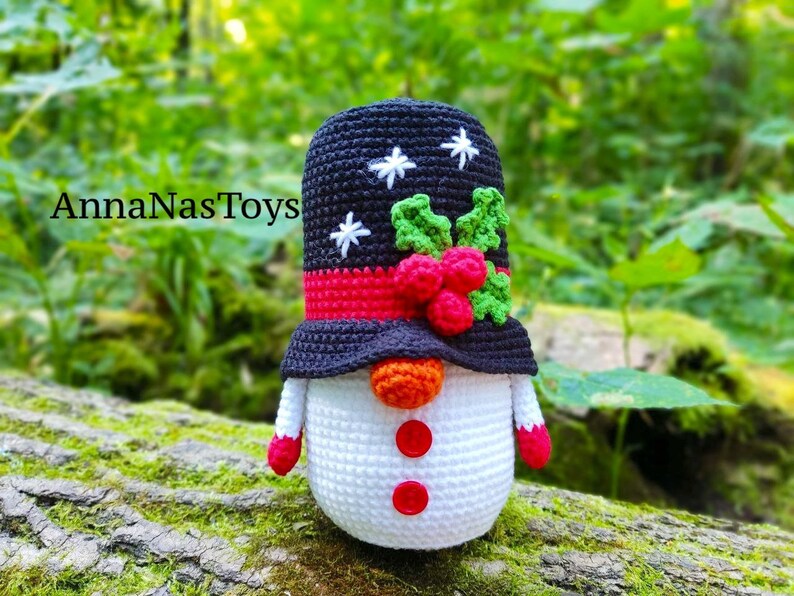 May include: A crocheted snowman ornament with a black hat, red and green holly, and white snowflakes. The snowman is wearing a red scarf and has an orange carrot nose and red buttons.