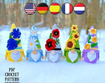 May include: Five crocheted gnomes with different flower designs on their hats. The gnomes are standing on a white surface. The gnomes are wearing yellow, blue, gray, green, and purple hats. The hats are decorated with flowers, including sunflowers, daisies, and daffodils. The gnomes are all smiling. The image includes the text "PDF CROCHET PATTERN ANNANASTOYS".
