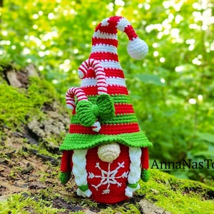 May include: A crocheted Christmas gnome with a red and white striped hat, green and white candy cane accents, and a white snowflake on the front. The gnome is sitting on a mossy log.