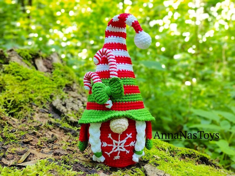 May include: A crocheted Christmas gnome with a red and white striped hat, green and white candy cane accents, and a white snowflake on the front. The gnome is sitting on a mossy log.