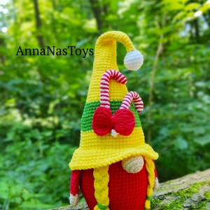 Puede incluir: Un gnomo de crochet con un cuerpo rojo, cabello amarillo y un sombrero amarillo y verde con un bast&oacute;n de caramelo y un lazo rojo. El gnomo est&aacute; sentado en un tronco de &aacute;rbol.