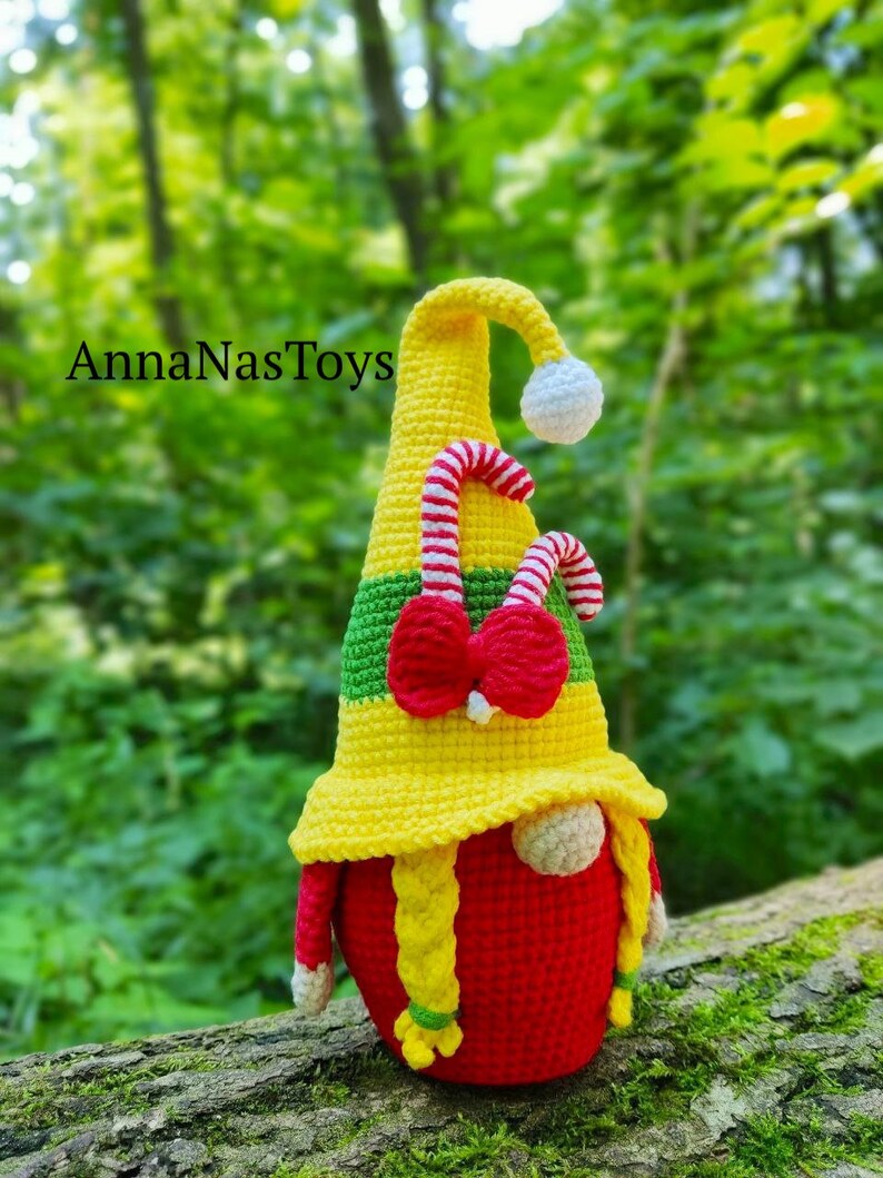 Puede incluir: Un gnomo de crochet con un cuerpo rojo, cabello amarillo y un sombrero amarillo y verde con un bast&oacute;n de caramelo y un lazo rojo. El gnomo est&aacute; sentado en un tronco de &aacute;rbol.