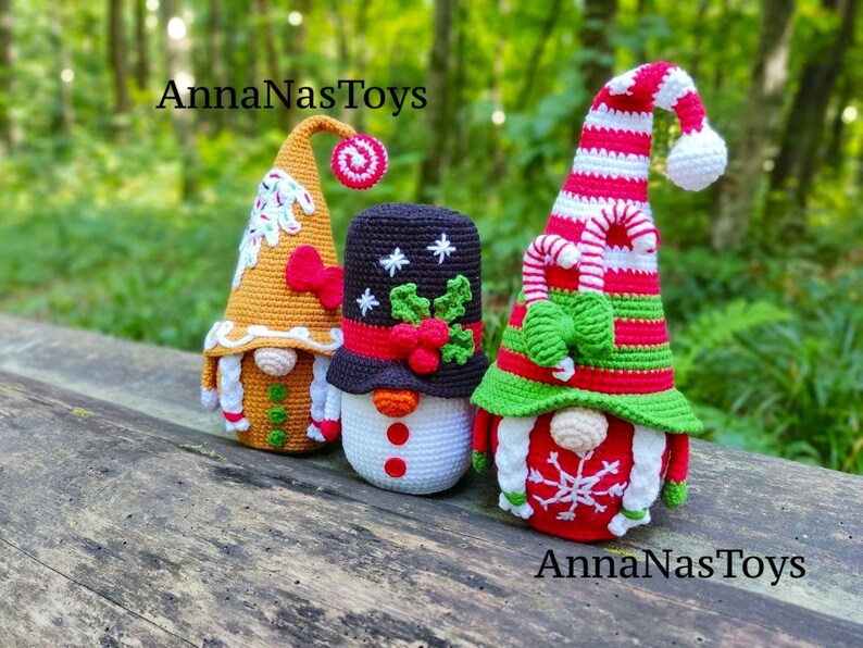 May include: Three crocheted Christmas gnomes, one gingerbread themed, one snowman themed, and one with a candy cane hat and green bow. The gnomes are sitting on a wooden surface with a green background. The text "AnnaNasToys" appears twice in the image.