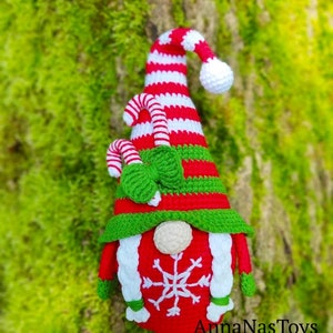 May include: A red and white striped gnome with a green hat and a candy cane. The gnome has a white beard and a snowflake on its chest. The gnome is standing in front of a green mossy background. The text "AnnaNasToys" is at the bottom of the image.