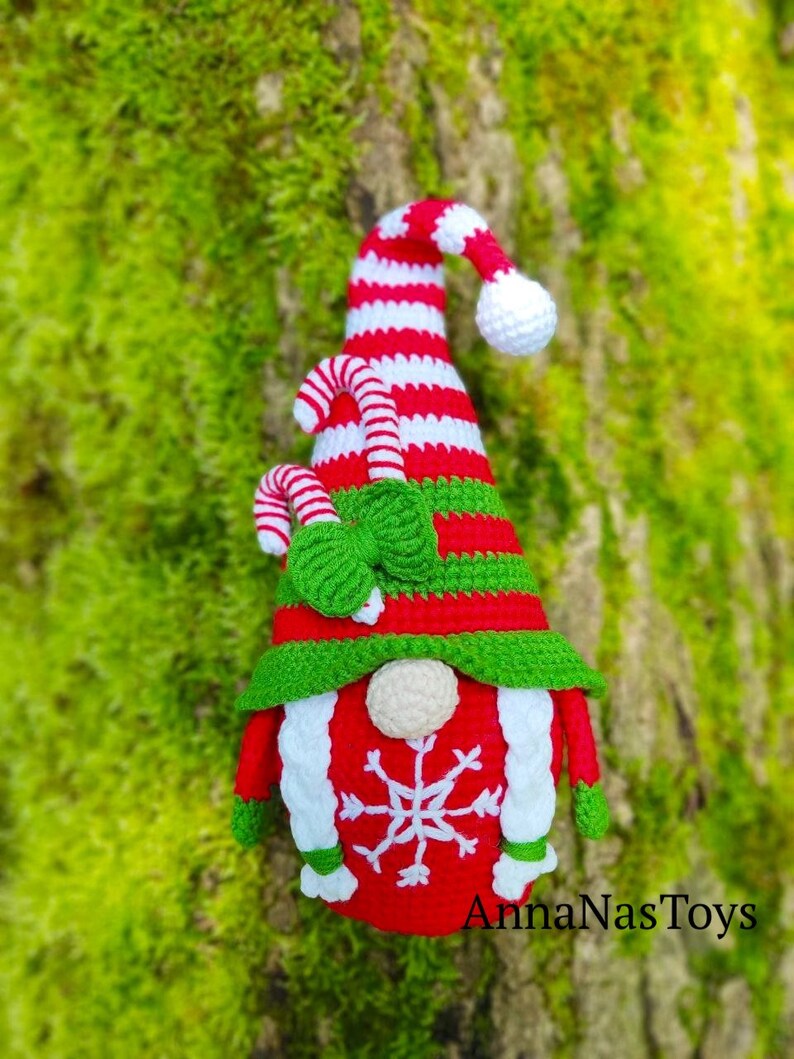 May include: A red and white striped gnome with a green hat and a candy cane. The gnome has a white beard and a snowflake on its chest. The gnome is standing in front of a green mossy background. The text "AnnaNasToys" is at the bottom of the image.