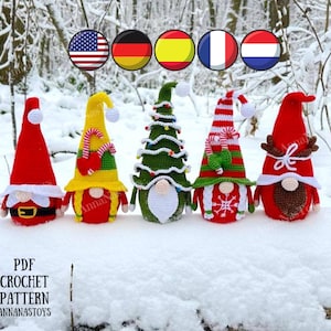 May include: Five crocheted gnomes in various colors and designs, including a Christmas tree gnome, a candy cane gnome, a yellow gnome with a candy cane, a red gnome with a white beard, and a red gnome with a brown beard. The gnomes are sitting on a snowy surface with a wooden log in the background. The image also includes flags of the United States, Germany, Spain, France, and the Netherlands.