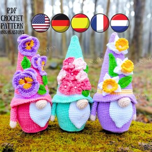 May include: Three crocheted gnomes with different flower designs on their hats. The gnome on the left has purple and yellow flowers, the middle gnome has pink flowers, and the gnome on the right has yellow and white flowers. The gnomes are sitting on a mossy log.