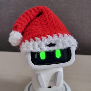 May include: A white AI robot with a black screen and green glowing eyes, wearing a red and white crocheted Santa hat. The robot is on a white base. The text "Amigurustitch" is at the bottom.