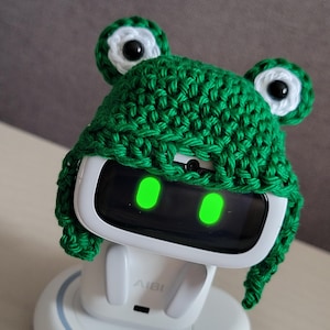 May include: A white AI robot with a green glowing screen, wearing a handmade green crochet frog hat with white and black eye details. The robot is on a white charging base. The text "Amigurustitch" is visible.