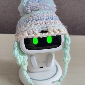 May include: A white AI robot with a crocheted hat. The hat is pastel-colored with a pom-pom on top and ear flaps. The robot's screen displays two green glowing eyes. The text "Amigurustitch" is at the bottom.