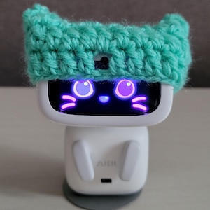 May include: A white AI robot with a screen displaying a cat face, wearing a turquoise crochet hat. The robot has a rounded body and ears. The text "Amigurustitch" is at the bottom.