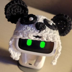 May include: A white and black crocheted panda hat with pearl accents is placed on a white electronic device with glowing green eyes. The hat has black ears and eyes. The text "Amigurustitch" is at the bottom.