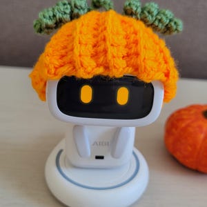 May include: A white robot toy with a black screen and orange eyes, wearing a crocheted orange pumpkin hat with green leaves. The robot sits on a white base with a blue ring. The text "Amigurustitch" is at the bottom.