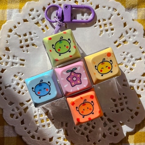 May include: A keychain featuring six colorful square charms with cartoon fruit faces. The charms are arranged in a cross shape and attached to a purple clasp. The background is a white doily on a yellow and white checkered surface.