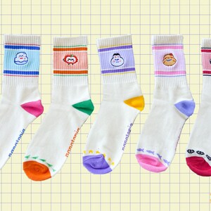 May include: Five pairs of white crew socks with colorful designs. Each sock features a different cartoon character on the cuff and a unique color scheme at the toe and heel. The socks have the word "nanatonia" printed on the side.