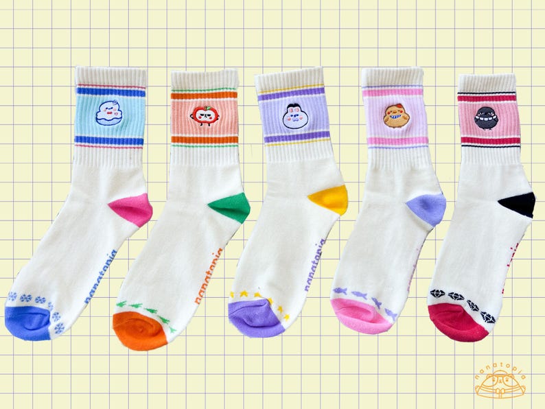 May include: Five pairs of white crew socks with colorful designs. Each sock features a different cartoon character on the cuff and a unique color scheme at the toe and heel. The socks have the word "nanatonia" printed on the side.