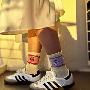May include: A pair of white sneakers with black stripes and a tan sole, worn with cream-colored socks. One sock has a red and yellow band with a cartoon character, the other has a purple band with a cartoon character.