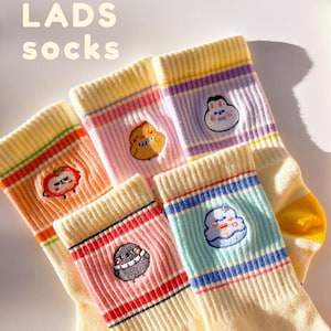 May include: A collection of cream-colored socks with colorful striped cuffs and embroidered cartoon characters. The socks feature an apple, a chicken, a bunny, a cupcake, and an egg. The text "LADS socks" is visible at the top.