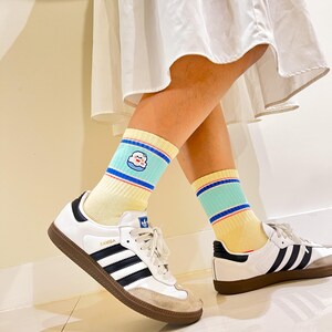 May include: A pair of white sneakers with black stripes and brown soles, paired with yellow socks featuring a cartoon design and blue and red stripes. The socks are worn with a white dress.
