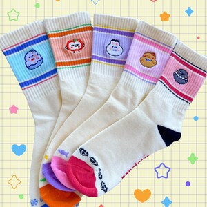 May include: Five pairs of cream-colored socks with colorful striped cuffs and embroidered cartoon characters. Each sock has a different design, including a blue cloud, a red apple, a purple dumpling, a yellow cookie, and a pink alien. The toes are multi-colored.
