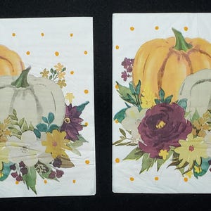 May include: Two square paper napkins featuring a white background with yellow polka dots. The design displays a watercolour illustration of a large orange pumpkin, a smaller white pumpkin, and a floral arrangement of dark purple and yellow flowers.