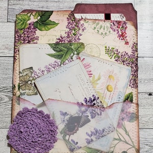May include: A decorative paper folder featuring a floral lilac print, containing several vintage-style postcards and ephemera. A small, round, purple crocheted doily is attached to the front pocket, which is made of translucent vellum paper.