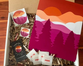 May include: A gift box filled with brown packing paper and a variety of pride-themed items. The box contains a print of a sunset over mountains in shades of pink, orange, and red, four stickers with the word "PRIDE" in red letters, two smaller stickers with a sunset over mountains design, and a black enamel pin with a sunset over mountains design.