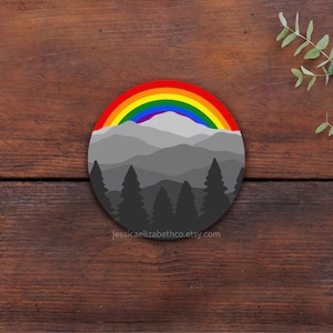 May include: A round coaster featuring a colorful rainbow over a mountain range and silhouetted trees. The coaster has a gray and black color scheme with a rainbow of colors. The text "jessicaelizabethco.etsy.com" is at the bottom.