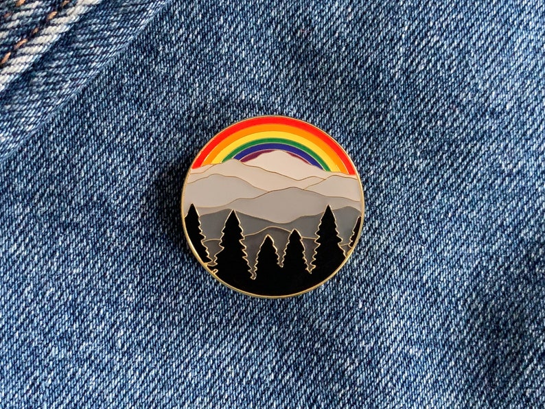 Op de afbeelding: Een emaille pin met een bergketen met een regenboog die over de toppen boogt. De bergen zijn grijs en de bomen zijn zwart.