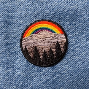 Può includere: Una toppa rotonda ricamata con un arcobaleno sopra una catena montuosa e una fila di alberi in primo piano. La toppa è nera con montagne viola e alberi verdi.