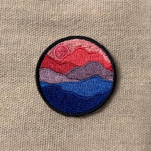 May include: A round embroidered patch with a sunset scene. The patch features a pink, red, purple, and blue color scheme with a layered mountain and wave design.