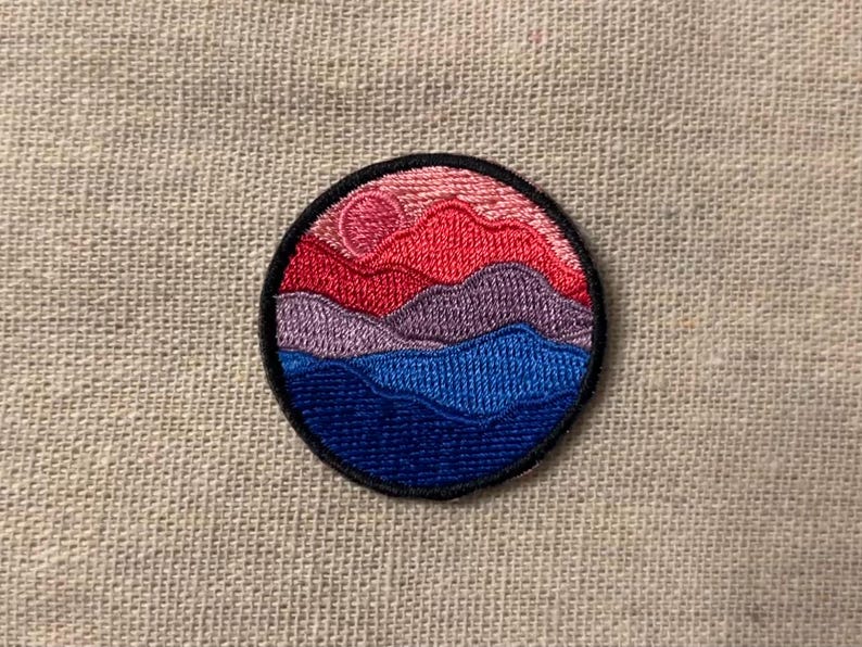 May include: A round embroidered patch with a sunset scene. The patch features a pink, red, purple, and blue color scheme with a layered mountain and wave design.
