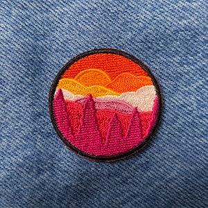 Può includere: Toppa ricamata con una scena di tramonto. La toppa presenta un tramonto rosa, arancione e giallo sopra una fila di alberi rosa. La toppa è circolare e ha un contorno nero.