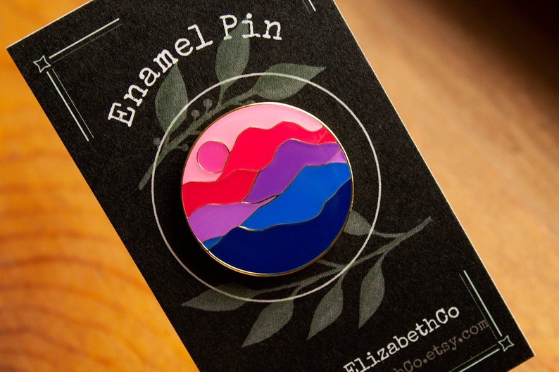 May include: A round enamel pin featuring a sunset over mountains in the colours of the bisexual pride flag. The pin is gold-toned and has a black background.