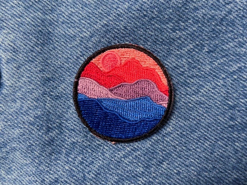 May include: A round embroidered patch with a sunset over mountains design. The patch features a pink and red sun, purple mountains, and blue water.