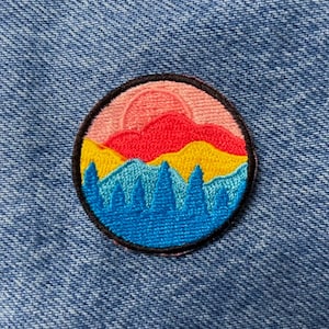 Könnte beinhalten: Ein runder gestickter Aufnäher mit einer Sonnenuntergangsszene. Der Aufnäher zeigt einen rosa und roten Himmel mit gelben und blauen Bergen im Vordergrund.