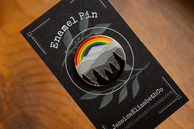 Op de afbeelding: Een ronde emaille pin met een regenboog boven een bergketen en een bos met bomen. De pin is zwart en goud met een witte en grijze bergketen en een regenboog met de kleuren rood, oranje, geel, groen, blauw, indigo en violet.