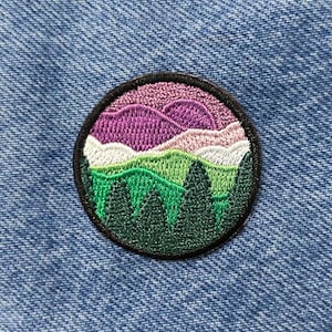 May include: A round embroidered patch with a landscape design. The patch features a purple, pink, white, green, and dark green color scheme. The design depicts a mountain range with trees in the foreground.