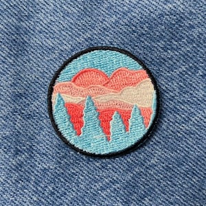 Peut inclure: Un patch rond brodé avec un fond bleu, des montagnes roses et blanches et un coucher de soleil rose. Le patch a une bordure noire.