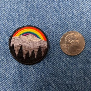 May include: Embroidered patch featuring a mountain range with a rainbow over it. The mountain range is purple and the rainbow is red, orange, yellow, green, blue, indigo, and violet. The patch is surrounded by a black border.