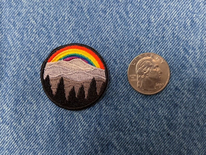 May include: Embroidered patch featuring a mountain range with a rainbow over it. The mountain range is purple and the rainbow is red, orange, yellow, green, blue, indigo, and violet. The patch is surrounded by a black border.