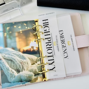 May include: A light pink leather binder with gold rings containing translucent dividers. One divider is labeled HIGH PRIOTIYY and another is labeled EMERGENCY with the text THE LIBRARY OF SAVINGS. A photo of a cozy blanket is visible on the left page.