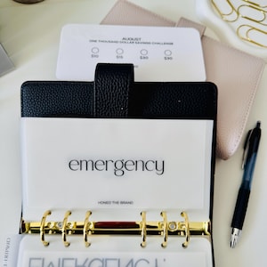 Könnte beinhalten: Ein schwarzer, texturierter Ordner mit einem goldenen Sechs-Ring-Mechanismus und durchscheinenden Trennblättern. Ein Trennblatt ist mit emergency und Honed The Brand beschriftet. Eine weiße Karte zeigt August One Thousand Dollar Savings Challenge.
